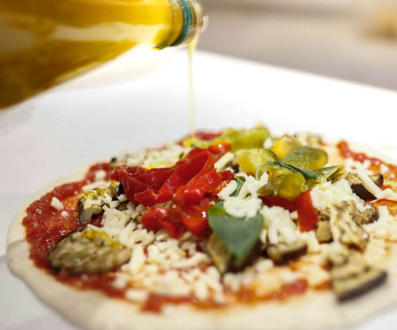 Un pizzaiolo verse de l'huile d'olives sur une pizza aux légumes